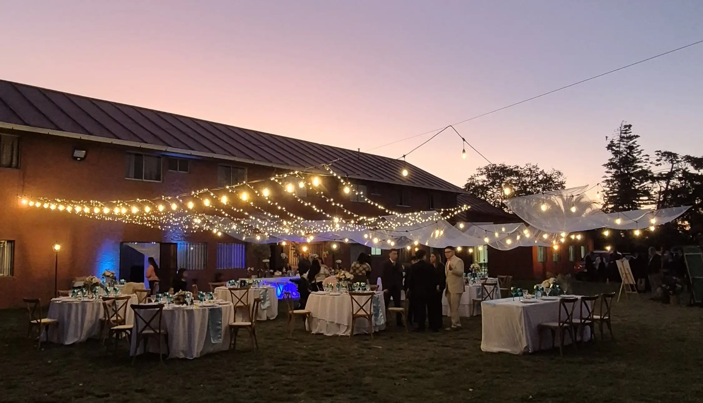 sobre-nosotros-espacio-mater Evento al atardecer en el jardín del centro de eventos Espacio Mater, con mesas decoradas elegantemente, luces colgantes cálidas y toldos blancos creando un ambiente acogedor y festivo.
